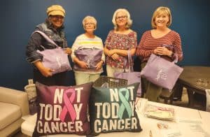 Four smiling women of various ages standing behind a table displaying "Not Today Cancer" throw pillows while holding "Nothing Ping" tote bags
